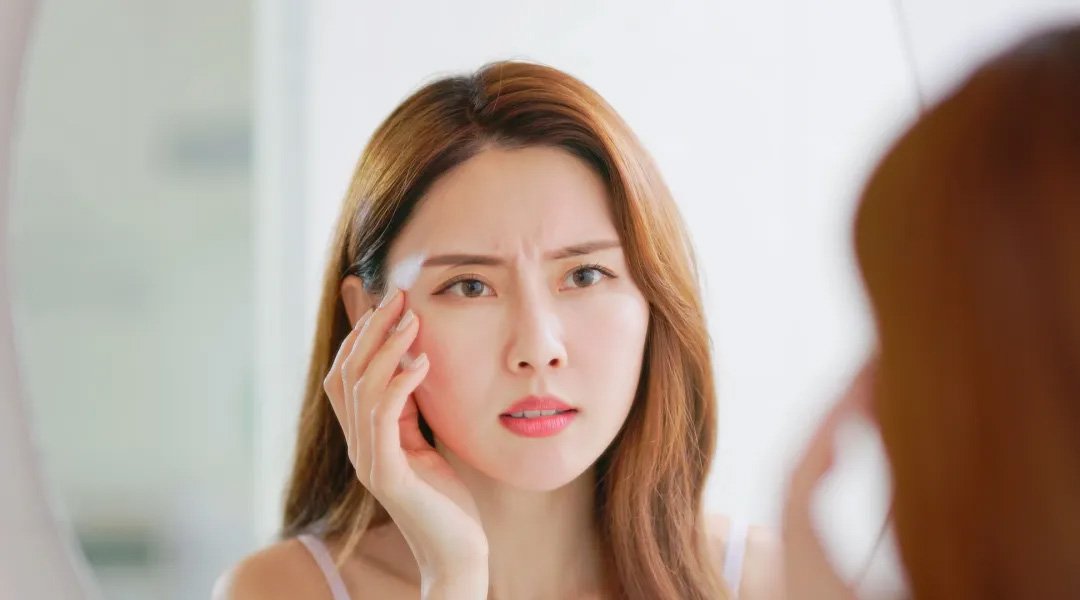 wanita di depan kaca dengan wajah khawatir karena makeup ngecrack